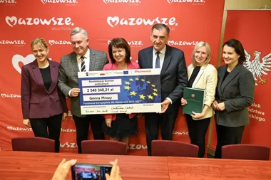 Grupa osób na tle czerwonej ścianki z logo Mazowsze trzyma duży czek symbolizujący dofinansowanie unijne dla Gminy Mrozy na rewitalizację terenów publicznych.